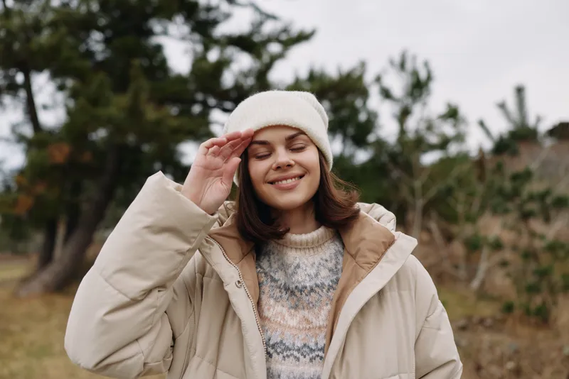 Woman in a cozy beige coat and beanie enjoying outdoor time in a park, smiling softly as she adjusts her hat, surrounded by trees and a calm autumn mood.