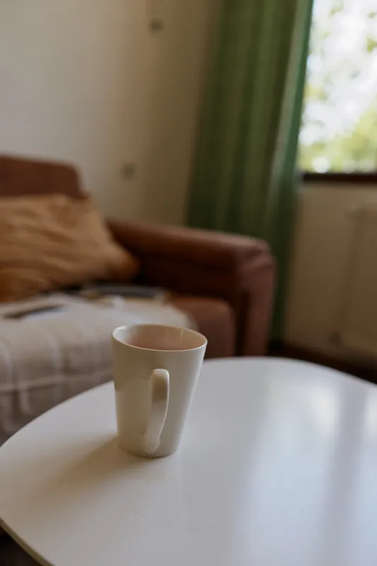 Cozy indoor scene features a white mug on a round table in a softly lit living room with a sofa, cushions and green curtains, evoking a peaceful morning ritual