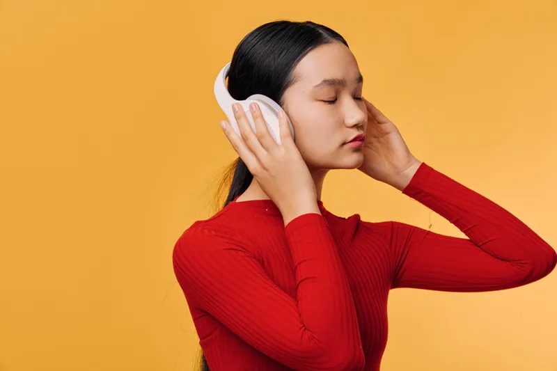 Woman wearing wireless headphones with eyes closed enjoying music for relaxation and entertainment against orange background. Asian female in red sweater listening to audio technology.