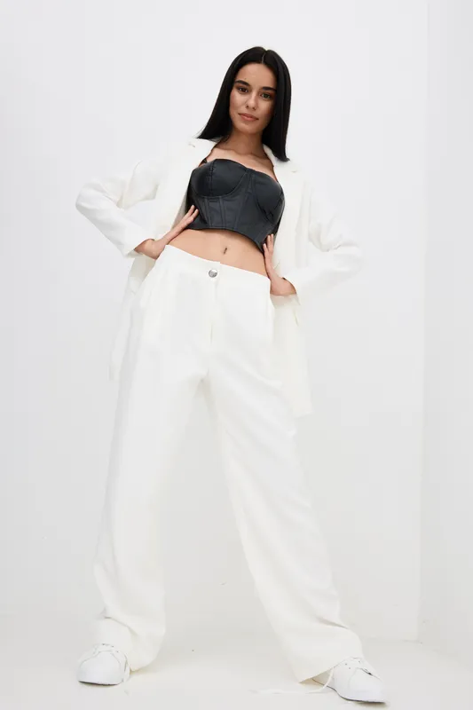 Young woman posing confidently in stylish white wide-leg pants, black crop top, and white blazer against a minimalist white background, showcasing modern fashion and elegance.