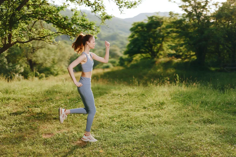 Young athletic woman jogging in a scenic green landscape, wearing activewear and expressing joy in sunny weather, promoting fitness and healthy lifestyle