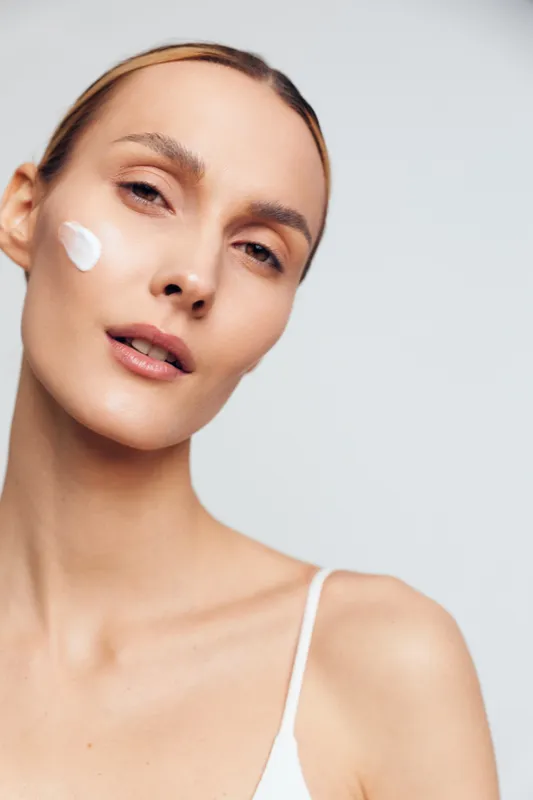 Young woman with clear skin applying white face cream on cheek, wearing white top. Close-up portrait on light background. Concept of skincare, beauty, health, and self-care routine.