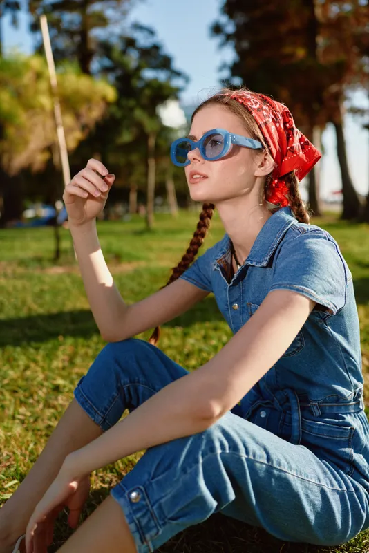 young woman sitting on grass wearing denim outfit and red headscarf enjoying sunny day outdoors with sunglasses and relaxed expression in natural park setting