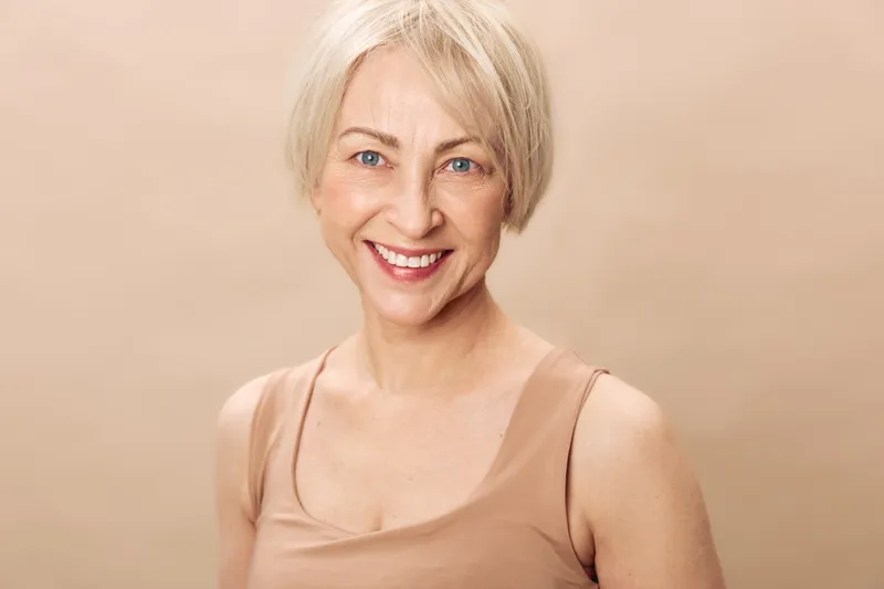 Smiling mature woman with short blonde hair wears beige tank top, looking at camera with bright blue eyes and natural makeup on soft neutral background. Concept for aging beauty and confidence.