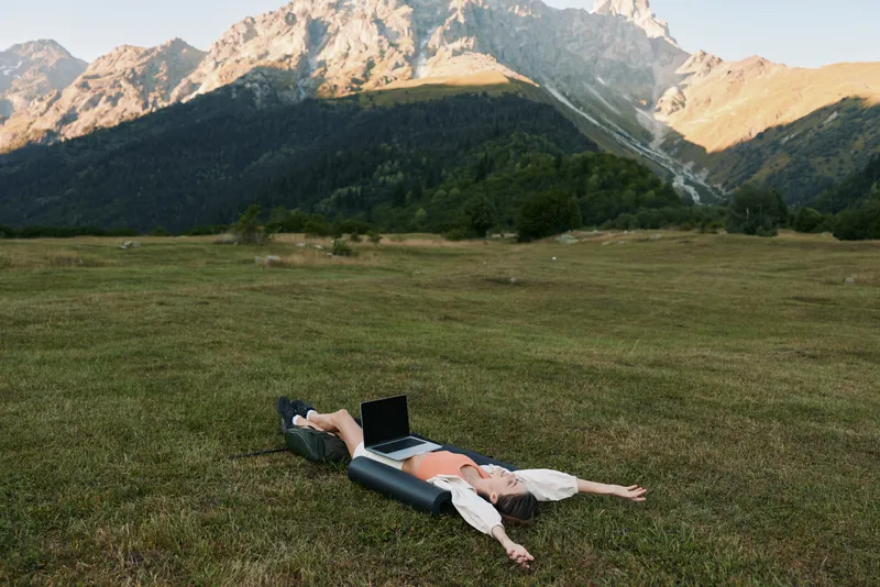 A relaxed professional lies on a green meadow with a laptop, embracing remote work in a scenic outdoor setting, enjoying fresh air, wide skies, and distant mountains framing a peaceful, productive