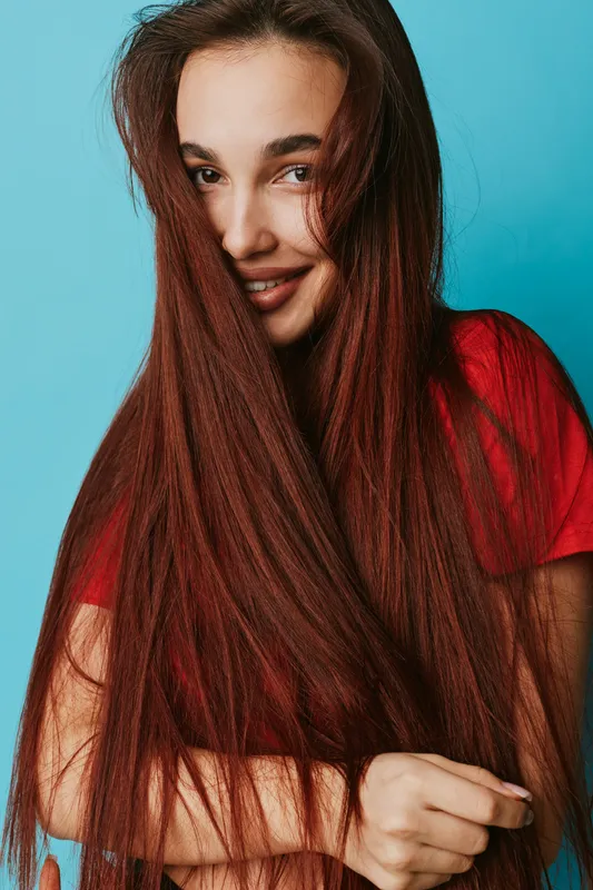 Woman with long red hair smiling and looking at camera against blue background. Casual portrait with natural makeup and bright red shirt in studio lighting for beauty and fashion.