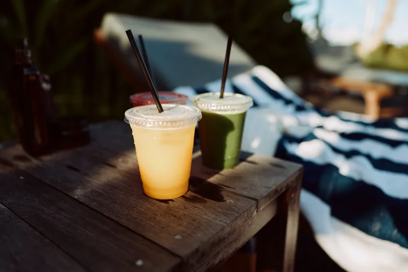 Bright outdoor scene with two colorful smoothies resting on a weathered wooden table, warm sunlight highlights the vibrant drinks, and a relaxed summertime atmosphere fills the background