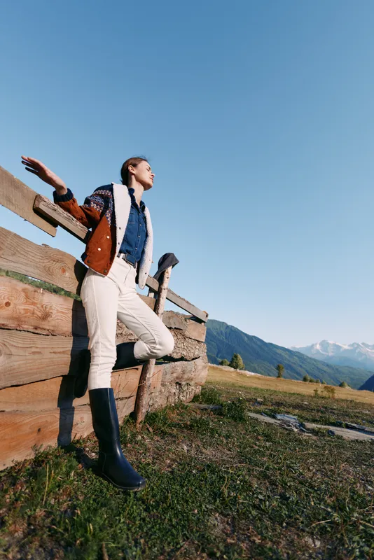 Woman leaning on wooden fence in mountains meadow wearing boots and jacket under clear blue sky in countryside, standing relaxed and enjoying scenic nature and open landscape.