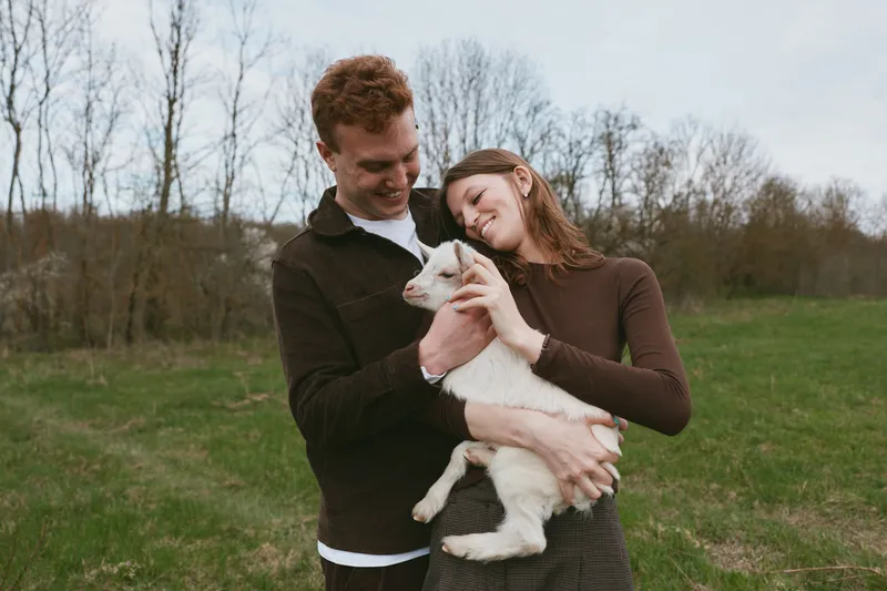 Smiling couple holding a white goat kid in a green field. Outdoor lifestyle moment showcasing bonding, care, and gentle affection in natural light. Soft shadows and warm tones. Gentle mood.