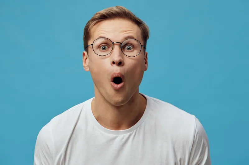 Surprised man with glasses wearing casual white T-shirt looking shocked in front of bright blue background. Studio portrait showing astonishment and curiosity. People emotion concept
