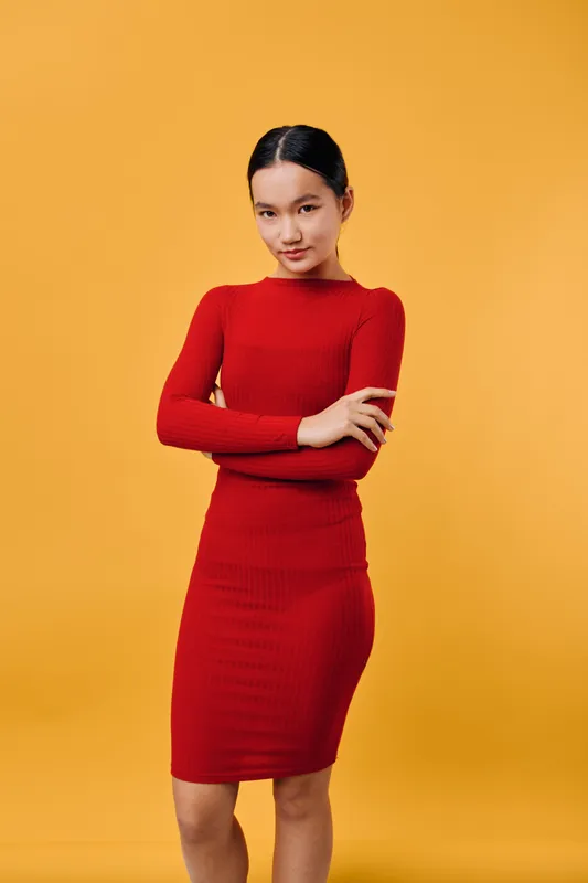 Young woman in red dress stands with crossed arms looking confidently at camera on yellow background. Female model with dark hair in neat style wears tight fitted outfit for fashion portrait.