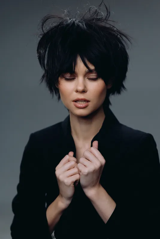 Young woman with tousled black hair wearing a black outfit expressing serene emotion against gray background