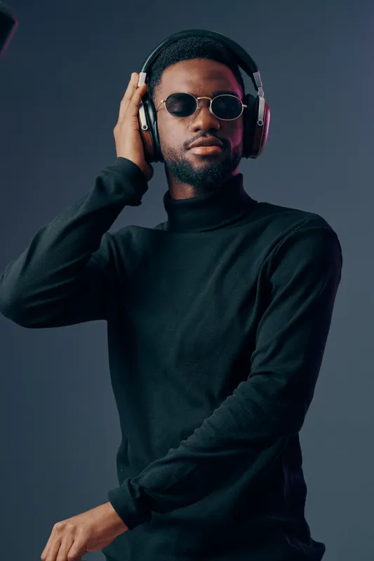 Stylish African man in black turtleneck and sunglasses enjoying music with headphones, conveying relaxation and confidence against a dark background
