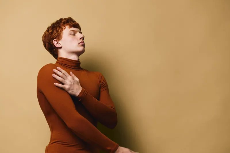 Young man with red hair wearing brown turtleneck posing with eyes closed and hand on chest against beige background in studio portrait.