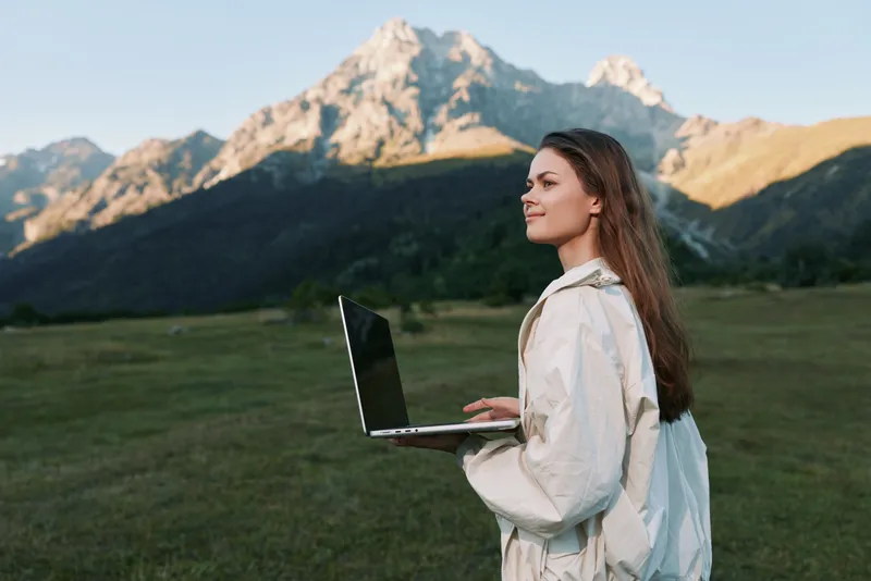 A calm woman stands in a lush meadow with a laptop, gazing toward distant mountains. Bright daylight highlights a focused, creative work moment, conveying productivity and outdoor inspiration.