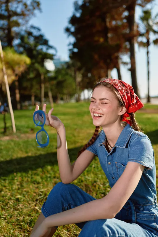 smiling woman in red headscarf holding blue sunglasses outdoors, happy young adult with braids, casual denim outfit, green park background on sunny day