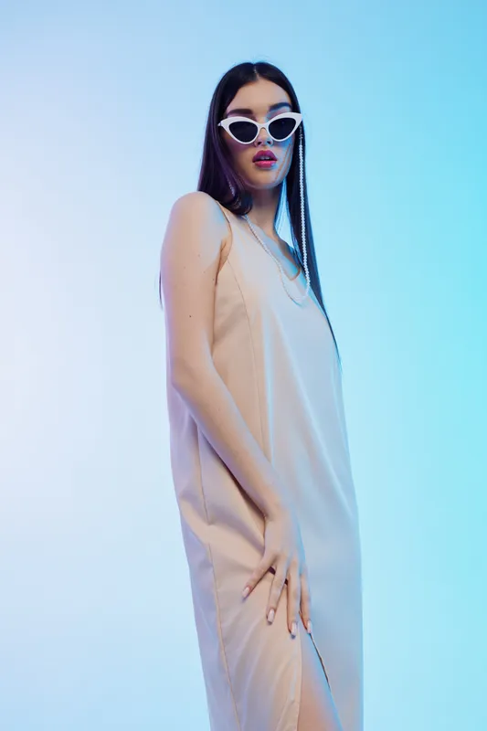 Fashionable woman in a beige dress and white sunglasses poses confidently against a blue gradient background. Concept of beauty, style, and modern fashion.