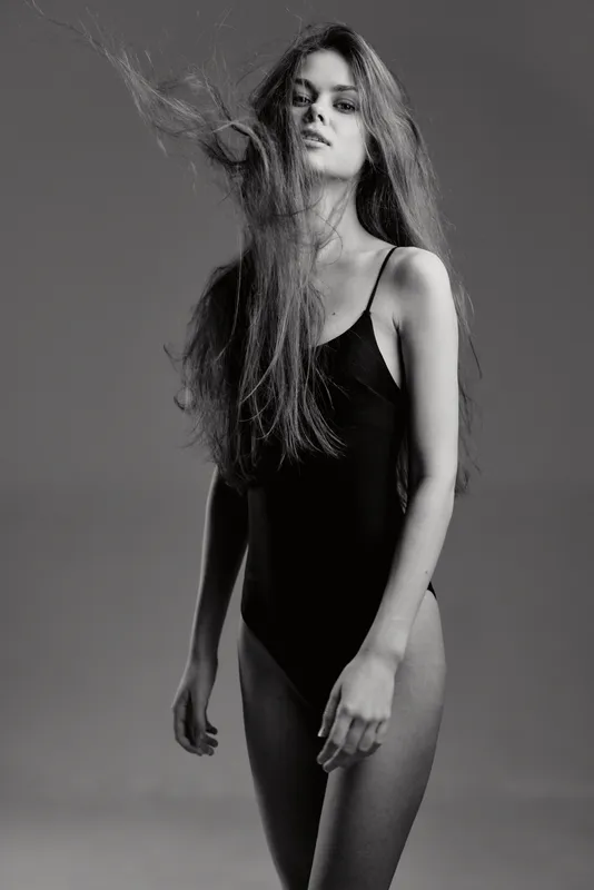 Black and white portrait of young woman in black bodysuit with flowing hair and serious expression on gray background