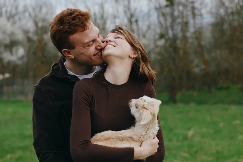 Young couple embraces outdoors while the woman holds a small white baby goat, sharing a tender moment in a natural setting with autumn colors and soft daylight.