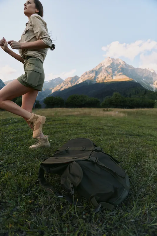A young woman in hiking attire leaps over a grassy meadow with a backpack, set against a dramatic mountain backdrop.