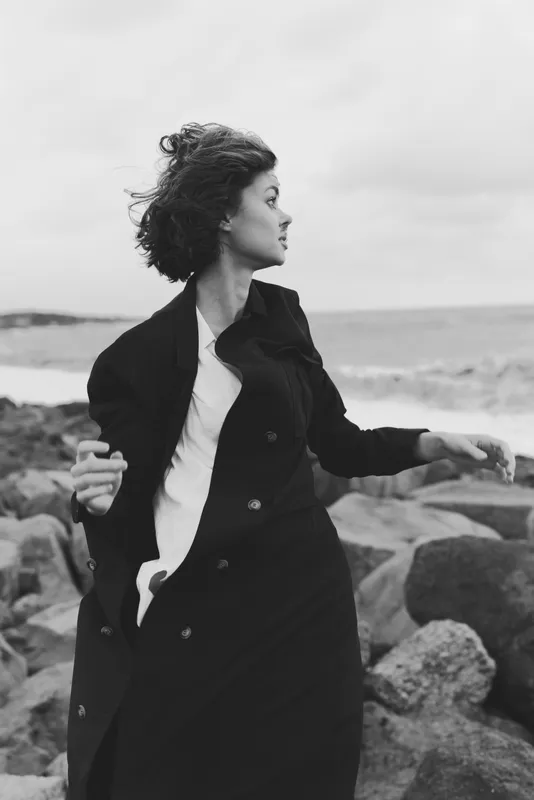 Woman in a long black coat stands on a windy coast by the sea, gazing toward the horizon, a moody monochrome scene with rugged rocks and ocean spray.