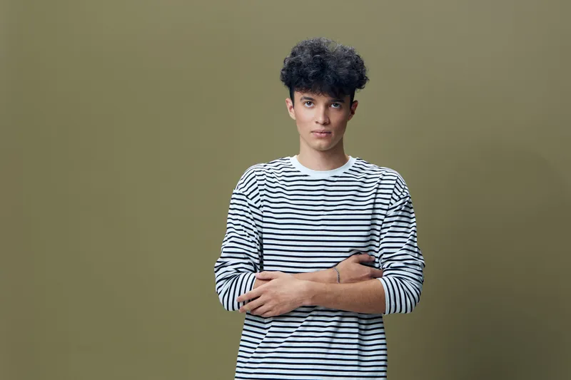 Young man with curly hair wearing a striped shirt standing with arms crossed against a muted olive background, calm and confident expression, studio portrait.