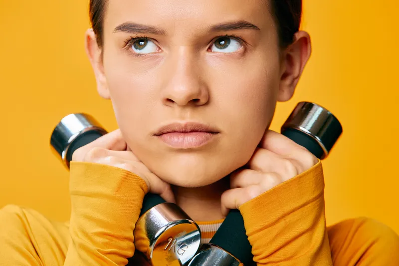 Woman fitness, weights and dumbbells lifted near shoulders in a studio portrait. Athletic female athlete in an orange top against a bright yellow background, focused expression, training and strength.