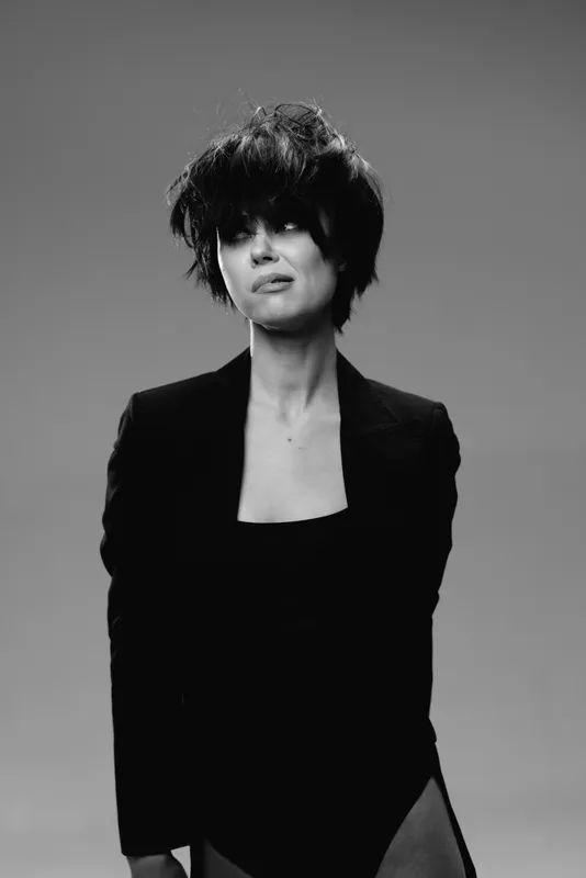Black and white portrait of a woman with short dark hair wearing a black outfit looking away with a thoughtful expression.