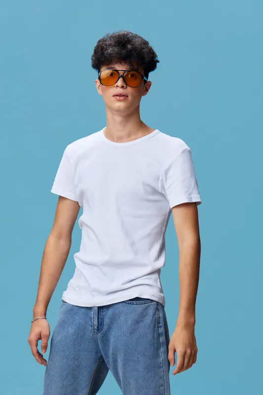 Young man wearing orange sunglasses and casual white t-shirt stands confidently against a solid blue background, showcasing a modern and relaxed style in daylight.