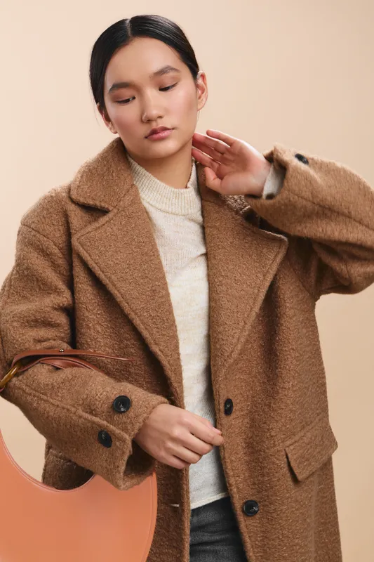 Asian woman wearing a warm camel coat poses in a neutral studio backdrop, her calm expression and poised posture conveying contemporary fashion, elegance, and quiet confidence.