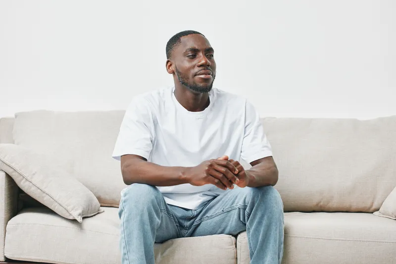 Smiling man sitting on a beige couch, wearing a white t shirt and jeans, conveys a relaxed atmosphere and a positive emotional state