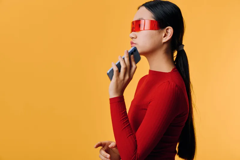 Woman with long hair wearing red futuristic glasses and holding a remote control standing against orange background in side profile pose with modern style and technology theme.