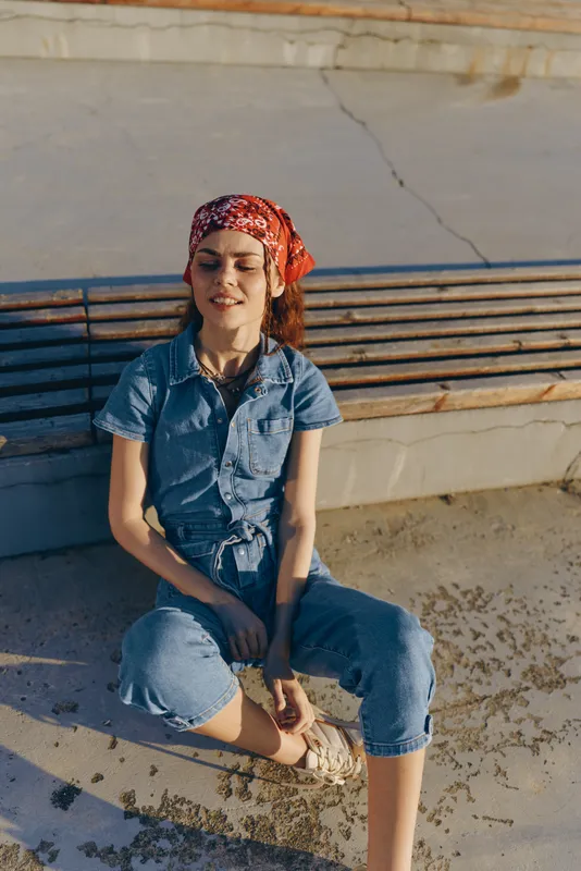 Lifestyle woman in modern boho-western denim street style wearing a red bandana sitting outdoors enjoying warm film color sunlight.