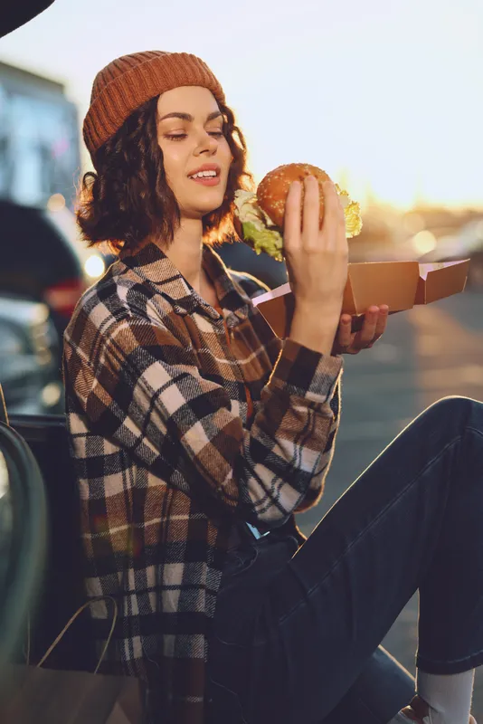 Woman burger outdoor golden car candid denim beanie picnic young woman eats a burger by a car at sunset, authenticity and candid lifestyle with golden hour glow, mindful living, emotional