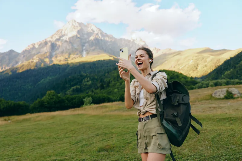 Young woman on a hiking trip takes a selfie in a mountain meadow, backpack ready, bright sky, cheerful mood amid vast nature and wildflowers sway nearby under blue sky.