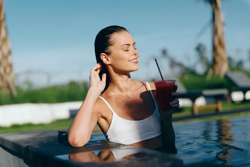 Cheerful woman relaxing in a sunlit pool, wearing a white swimsuit, holding a cold drink with a straw, eyes closed in contentment, warm breeze, lush outdoor setting, vibrant natural colors, carefree