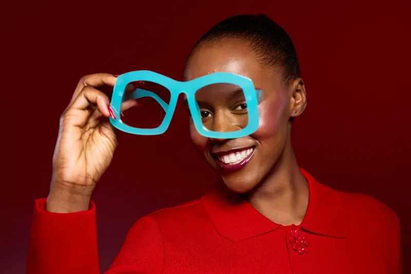 Happy African woman with dark skin holding oversized blue glasses in front of her face. She wears a red sweater and smiles against a dark red background. Concept of joy and fashion.
