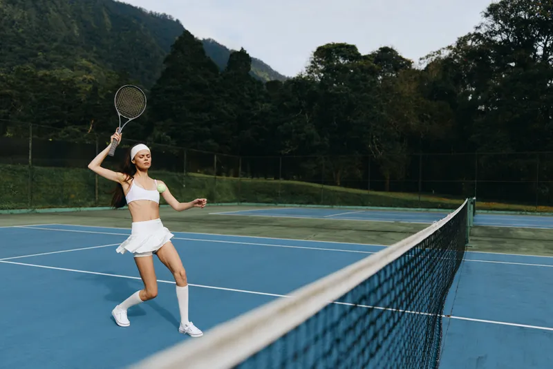 Energetic young female tennis player in a white athletic outfit, joyfully hitting the ball on a sunny day, showcasing fitness and sport lifestyle on a beautifully landscaped court