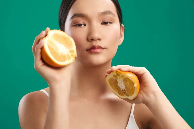 Asian woman holds sliced lemon halves near her face, posed against a vibrant green backdrop. The bright citrus contrast enhances her calm expression, creating a fresh, playful portrait with a clean,