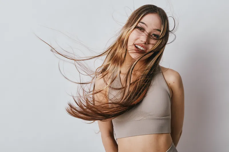 Young woman with long flowing hair and casual top smiling while posing against plain light background. Natural beauty and motion captured with joyful expression and loose hair strands.