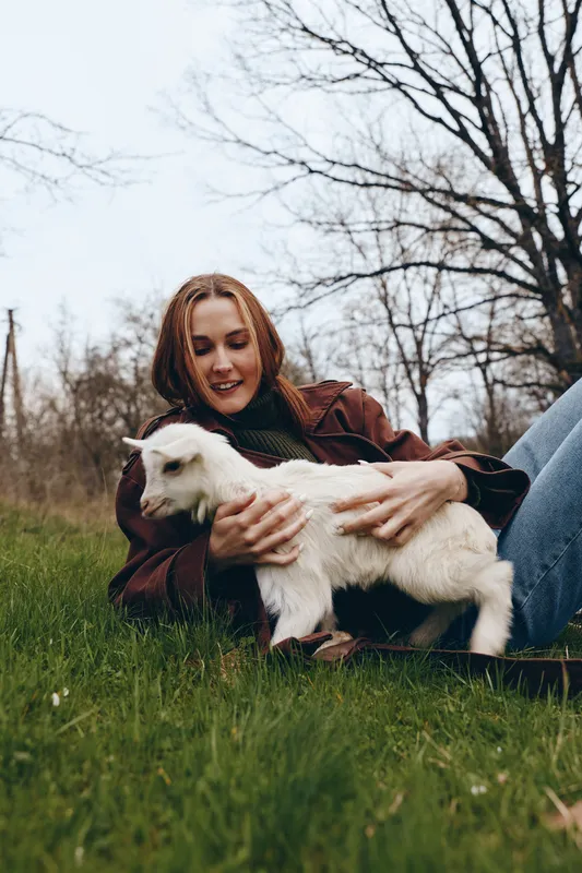 A woman outdoors shares a gentle moment with a playful goat kid on a grassy field, conveying a relaxed lifestyle and affectionate bond between human and animal.