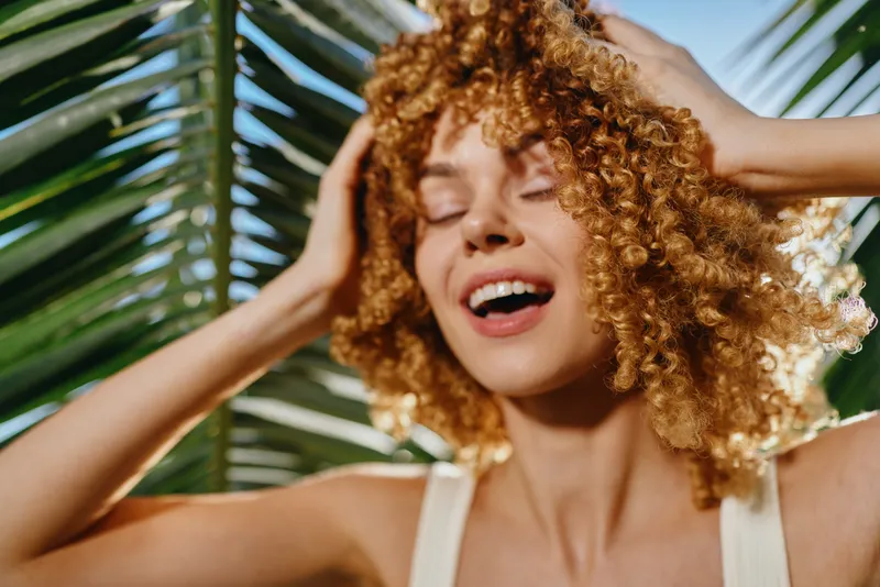 Smiling woman with curly hair enjoying sunlight, tropical palm leaves background, summer vibe, natural beauty, happy expression.