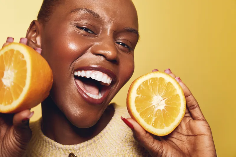 Happy smiling black woman holds two orange halves near face on yellow background. African ethnicity female with dark skin shows fresh fruit concept for health, nutrition, and vitamin C.