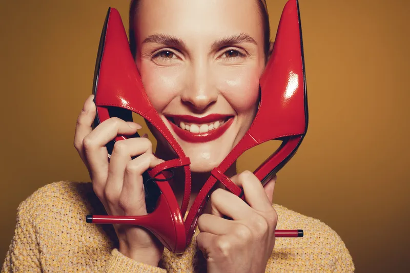 Smiling woman holds red high heel shoes near face, wearing yellow sweater. Close-up portrait on brown background. Concept of fashion, style, and happiness with bright makeup and accessories.