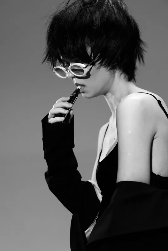 Black and white portrait of a thoughtful woman wearing sunglasses and a dark outfit in a minimalist setting.