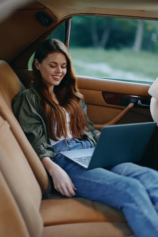 Young woman working on a laptop inside a car, wearing casual clothes, smiling, with a natural green background promoting remote work and digital nomad lifestyle.