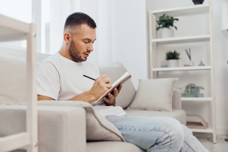 Focused man writing in a notebook, dressed casually in a white t-shirt, seated in a cozy living room, surrounded by indoor plants and natural light.