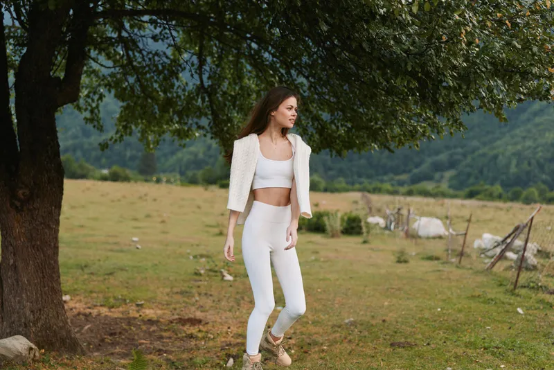 Young woman in white outfit walks near a tree in a green meadow with mountains in the background, enjoying a peaceful outdoor moment.