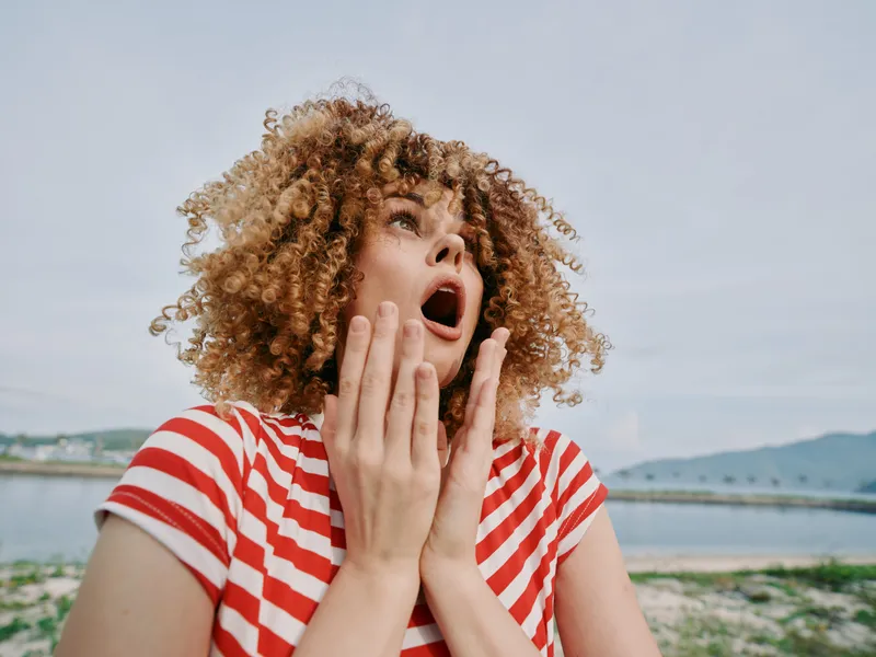 Main focus on a surprised woman with curly hair wearing a red and white striped shirt, hands near her face, expressive reaction outdoors by a calm waterfront.