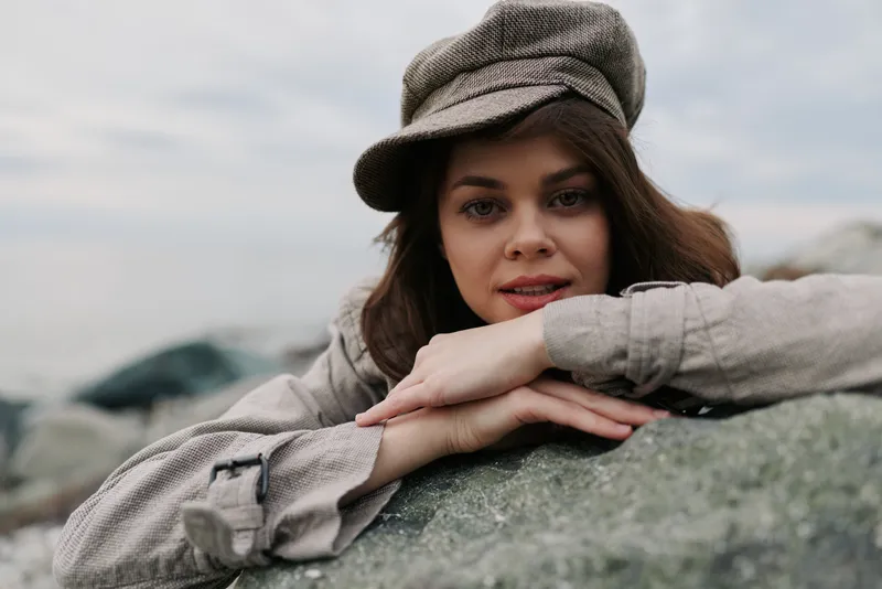 Young woman in a casual hat rests on rocks outdoors, looking at camera with soft expression. Natural light highlights her face and relaxed mood in a cool outdoor setting.